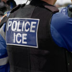 Immigration and Customs Enforcement - Close-up of POLICE ICE marking on the back of a hi-visibility stab proof vest worn by a trio of police officers at the scene of an incident.