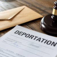 A deportation document lies on a wooden table with a brown envelope and a judge's gavel, conveying a serious and legal tone.