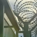 Close-up of a tall metal fence topped with coiled barbed wire under a cloudy sky. The scene evokes a sense of security and confinement.