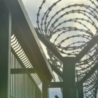 Close-up of a tall metal fence topped with coiled barbed wire under a cloudy sky. The scene evokes a sense of security and confinement. Close-up of a tall metal fence topped with coiled barbed wire under a cloudy sky. The scene evokes a sense of security and confinement.