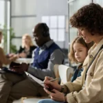 A woman with curly hair sits with a young girl in a busy waiting room, filling out a form. Adults in the background are engaged in conversations. Casual atmosphere.